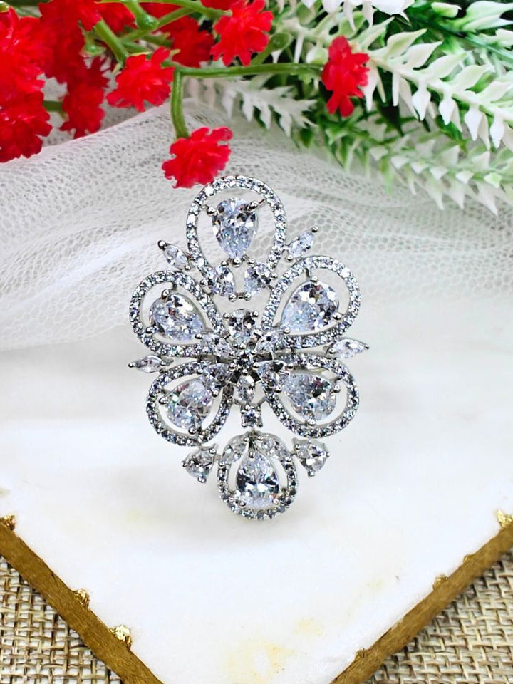 The Melissa White cocktail Ring is a sparkling silver ring with an intricate floral design, featuring White teardrop gemstones and clear American Diamonds, elegantly displayed on a white marble slab with red and white flowers in the background.