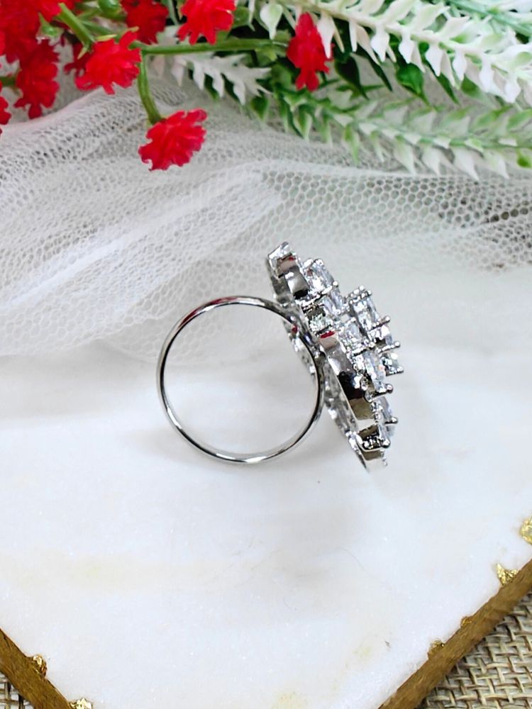 The Melissa White cocktail Ring is a sparkling silver ring with an intricate floral design, featuring White teardrop gemstones and clear American Diamonds, elegantly displayed on a white marble slab with red and white flowers in the background.