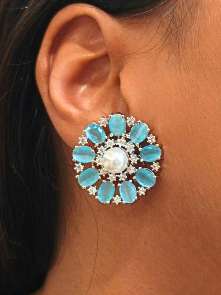 A close-up of a womans ear wearing the Mint Blue Blossom stud Earrings—large, circular Mint Blue earrings with a pearl at the center, colorful accents, and a border of sparkling American Diamonds. The stud earrings give a flower earrings feel. 