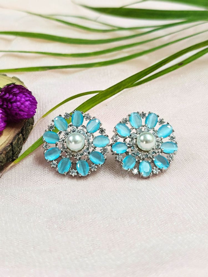 The Mint Blue Blossom Earrings showcase a central white pearl encircled by alternating Mint Blue stones and American diamonds, elegantly displayed on soft pink fabric with green leaves and a purple flower.