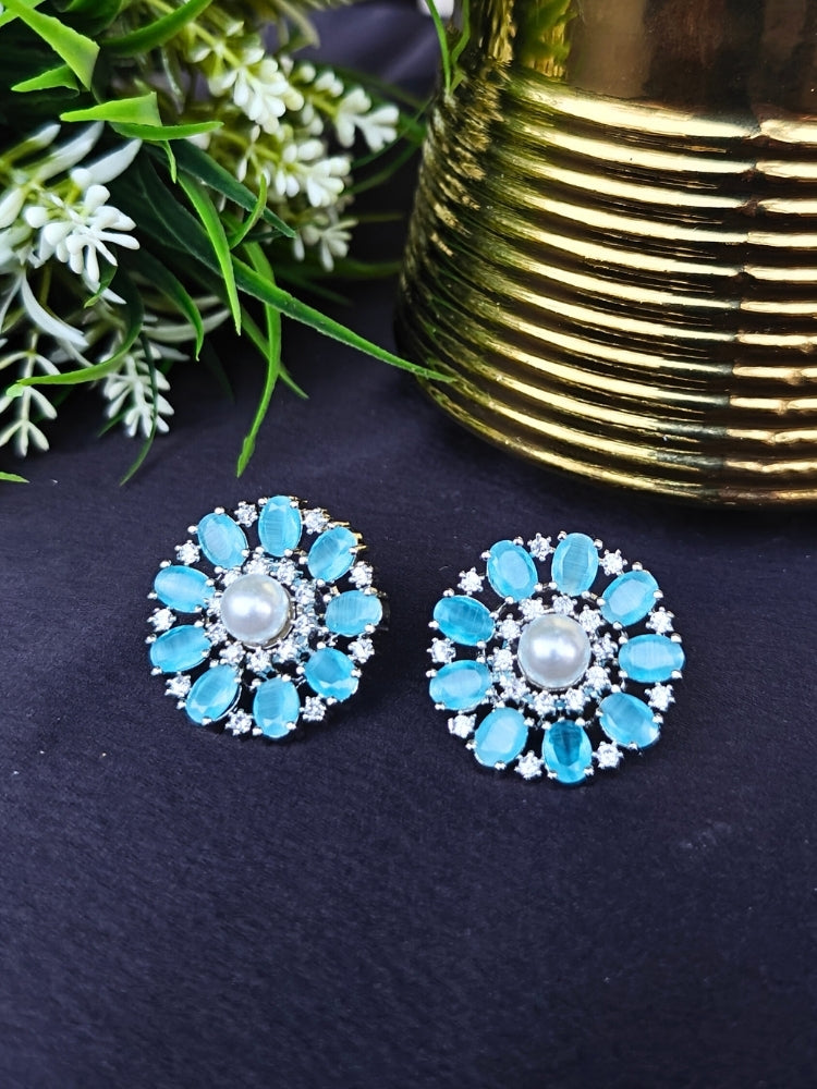 The Mint Blue Blossom Earrings showcase a central white pearl encircled by alternating Mint Blue stones and American diamonds, elegantly displayed on Black fabric with green leaves and a Golden accent.