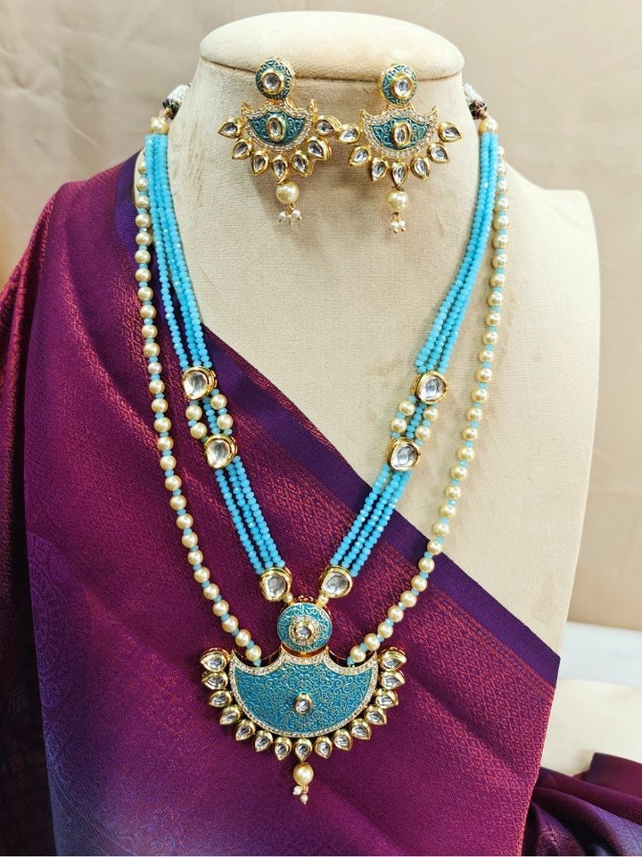 A mannequin in a deep maroon sari showcases the Mint Blue Zareen Necklace Set, featuring gold and turquoise tones with pearl accents, matching earrings, intricate designs, american diamonds, and elegant Kundan detailing.