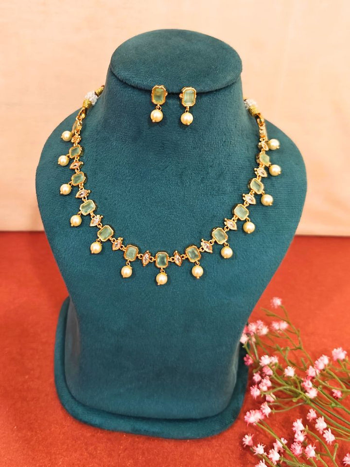The Mint Green Amelia Choker Necklace Set features colored stones and dangling pearl-like beads, displayed on a teal velvet stand, with matching stud earrings above and pink flowers beside the stand.