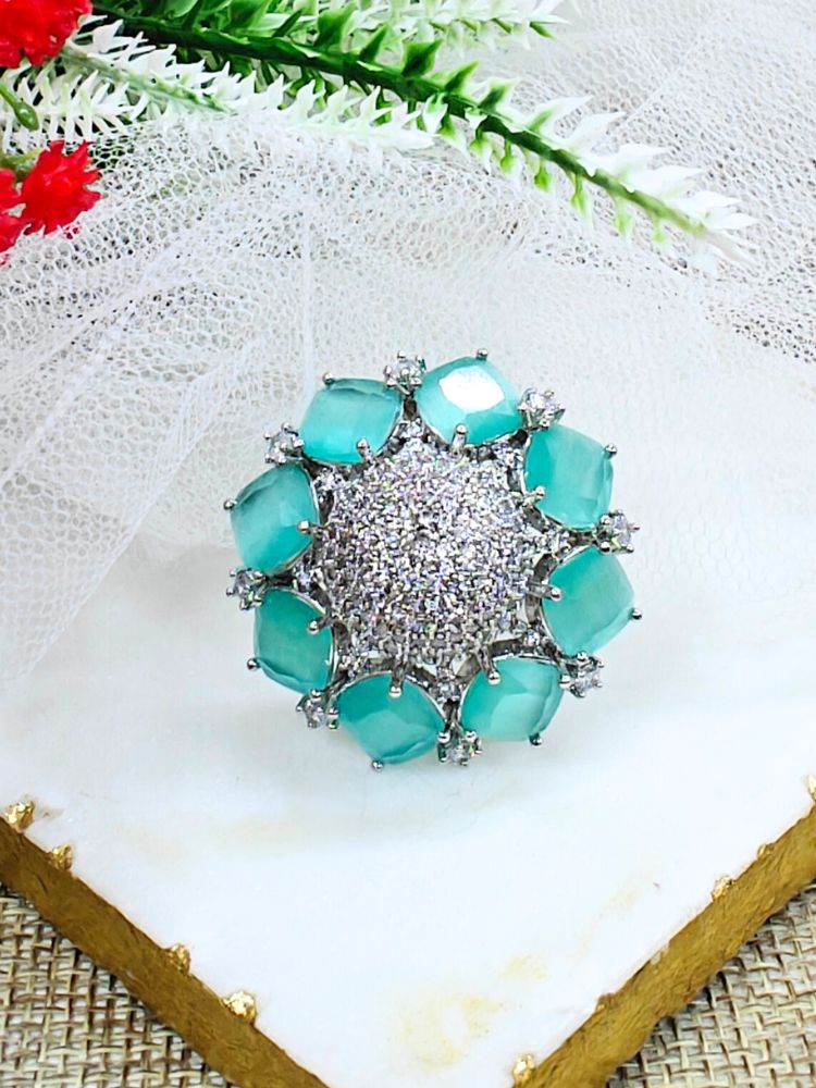 The Mint Green Aster Ring is a round silver piece with a sparkling, round-shaped center made with white American diamonds, surrounded by petal shaped Mint Green gemstones, along with an adjustable band. It’s shown on a textured surface with white netting and green leaves in the background.