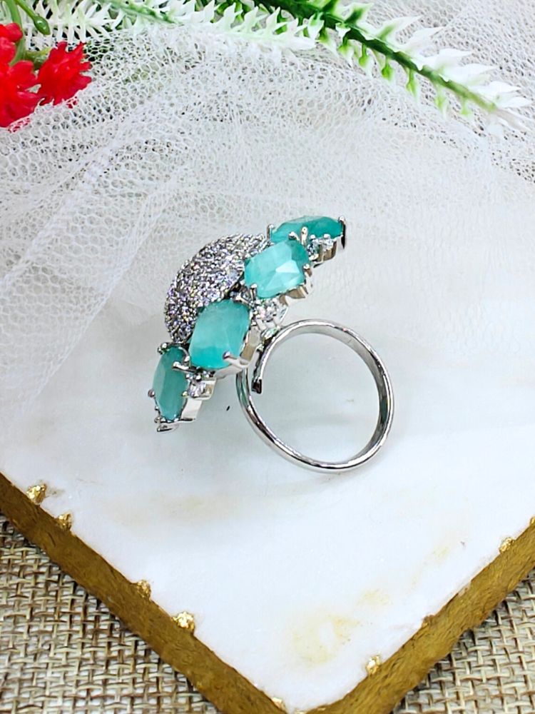 The Mint Green Aster Ring is a round silver piece with a sparkling, round-shaped center made with white American diamonds, surrounded by petal shaped Mint Green gemstones, along with an adjustable band. It’s shown on a textured surface with white netting and green leaves in the background.