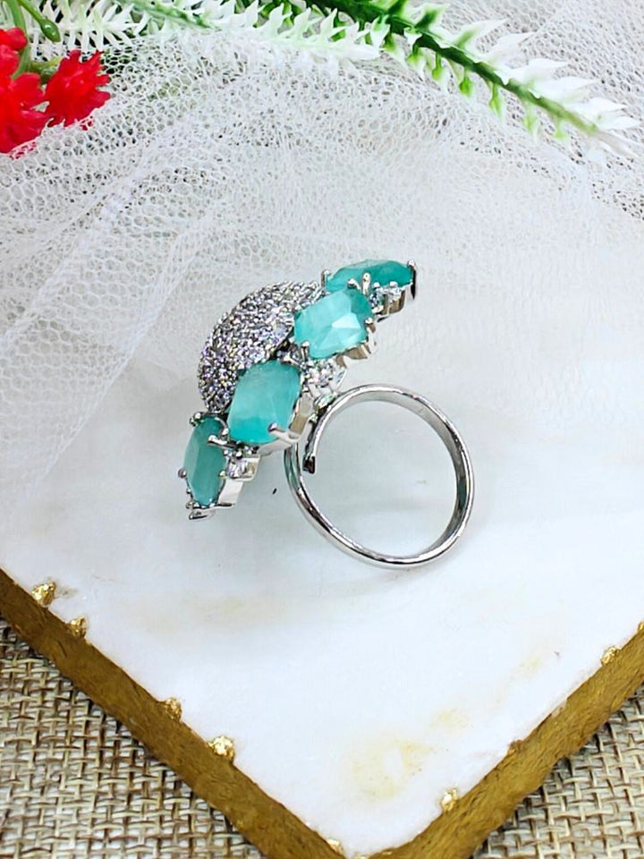 The Mint Green Aster Ring is a round silver piece with a sparkling, round-shaped center made with white American diamonds, surrounded by petal shaped Mint Green gemstones, along with an adjustable band. It’s shown on a textured surface with white netting and green leaves in the background.