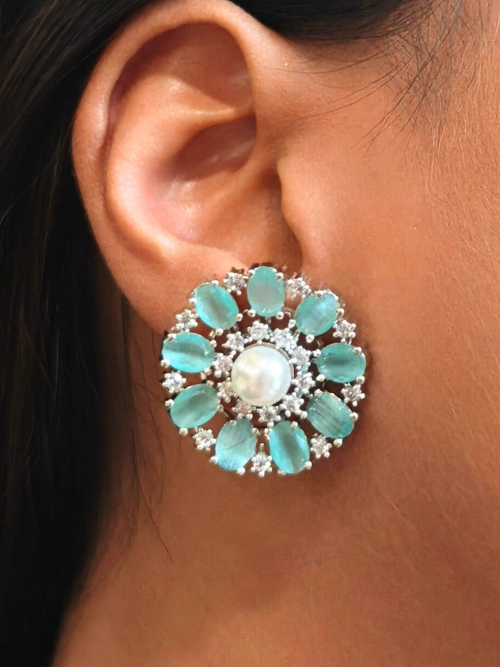 A close-up of a womans ear wearing the Mint Green Blossom stud Earrings—large, circular Mint Green earrings with a pearl at the center, colorful accents, and a border of sparkling American Diamonds. The stud earrings give a flower earrings feel. 