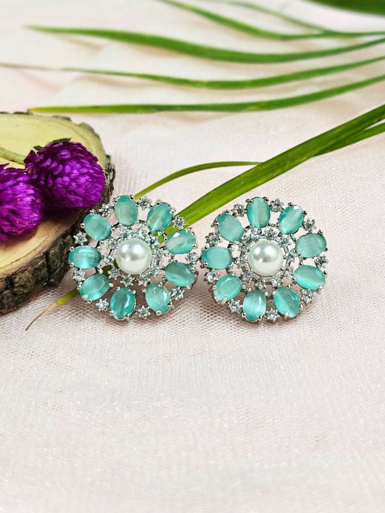 The Mint Green Blossom Earrings showcase a central white pearl encircled by alternating Mint Green stones and American diamonds, elegantly displayed on soft pink fabric with green leaves and a purple flower.