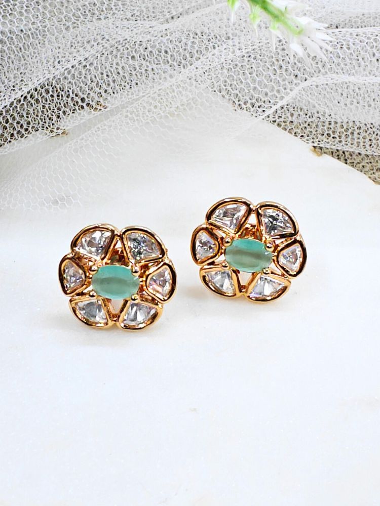 The Mint Green Daffodil stud Earrings feature gold flower-shaped brass settings with clear American Diamond petals and a mint green stone center, displayed on a white surface with sheer white fabric in the background.