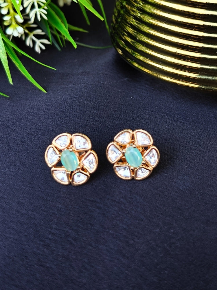 The Mint Green Daffodil stud Earrings feature gold flower-shaped brass settings with clear American Diamond petals and a mint green stone center, displayed on a Black surface with Golden Accent in the background.