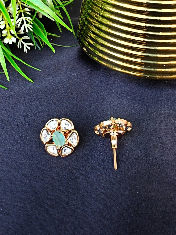 The Mint Green Daffodil stud Earrings feature gold flower-shaped brass settings with clear American Diamond petals and a mint green stone center, displayed on a Black surface with Golden Accent in the background.