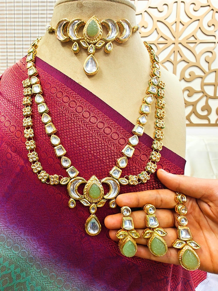 The Mint Green Nadira Necklace Set, features a tripple layered necklace in which there is a seperate choker made with carved stone in the center, along with kunda and pearl detailing on the side. The second necklace is a 2-layered necklace made with with curved stone in the center, along with kundan detailing all along the necklace. It comes with matching earrings. It is displayed on a mannequin in a maroon and teal saree; a hand holds the earrings.