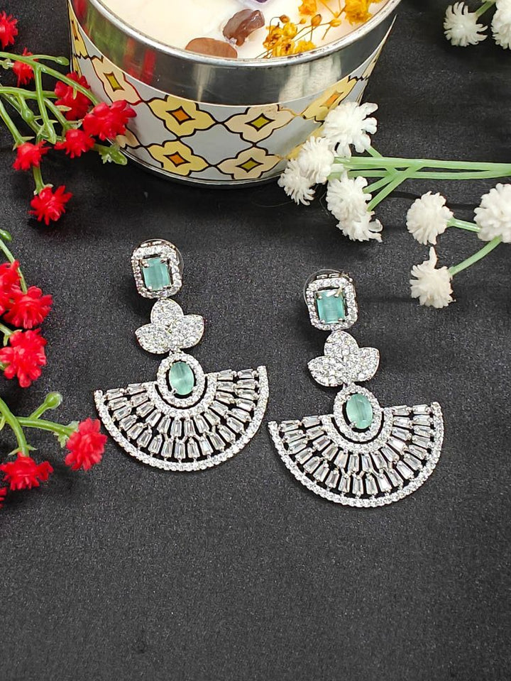 The Mint Green Nora Earrings feature ornate fan-shaped designs with American Diamonds, displayed on a black surface near red and white flowers and a decorative bowl with floating elements.