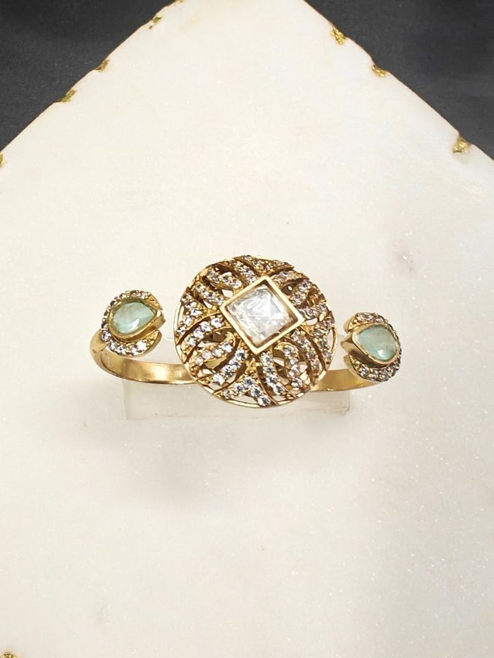 The Mint Green Octavia Ring features a gold band with a round, ornate centerpiece set with a clear American Diamond and two smaller Mint Green gemstones on the side, displayed on a white surface with gold trim. Multi-finger ring, great for cocktail parties. 