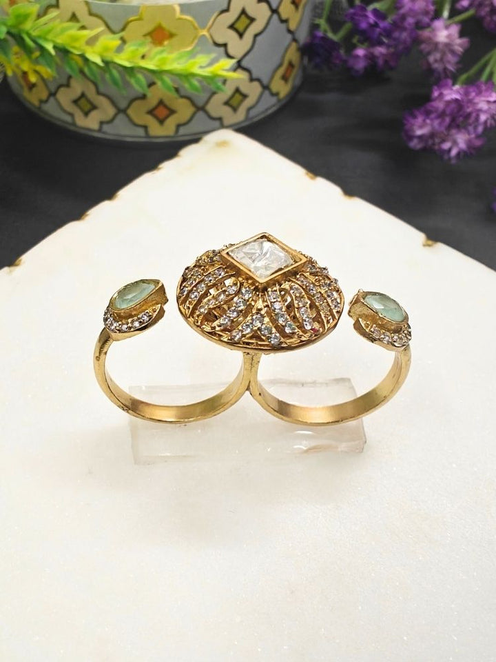 The Mint Green Octavia Ring features a gold band with a round, ornate centerpiece set with a clear American Diamond and two smaller Mint Green gemstones on the side, displayed on a white surface with gold trim. Multi-finger ring, great for cocktail parties.
