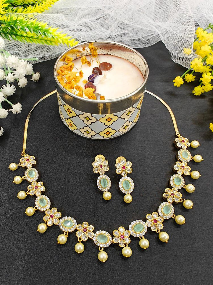 The Mint Green Samrina American Diamond Necklace Set, featuring American diamonds, green stones, pearls, and matching earrings, is displayed on a black surface next to a patterned candle jar, surrounded by white and yellow flowers.