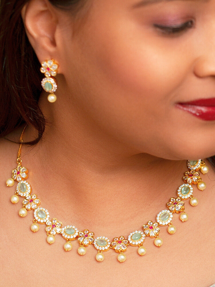The Mint Green Samrina American Diamond Necklace Set, featuring American diamonds, green stones, pearls, and matching earrings, is displayed on a black surface next to a patterned candle jar, surrounded by white and yellow flowers.