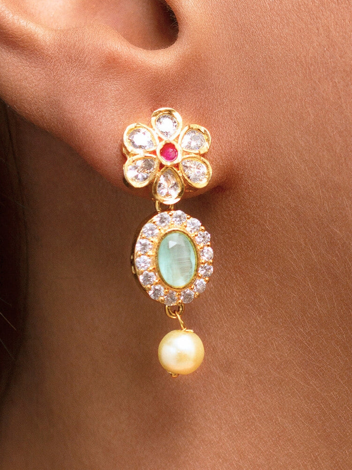 The Mint Green Samrina American Diamond Necklace Set, featuring American diamonds, green stones, pearls, and matching earrings, is displayed on a black surface next to a patterned candle jar, surrounded by white and yellow flowers.
