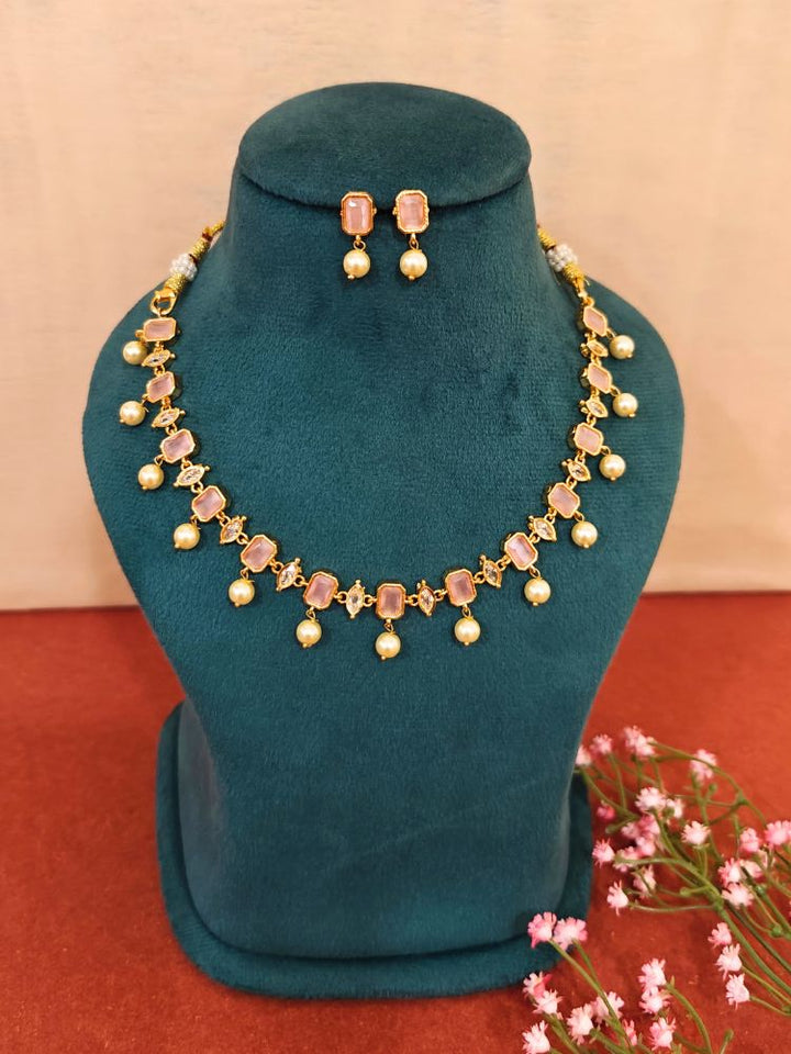 The Mint Pink Amelia Choker Necklace Set features colored stones and dangling pearl-like beads, displayed on a teal velvet stand, with matching stud earrings above and pink flowers beside the stand.