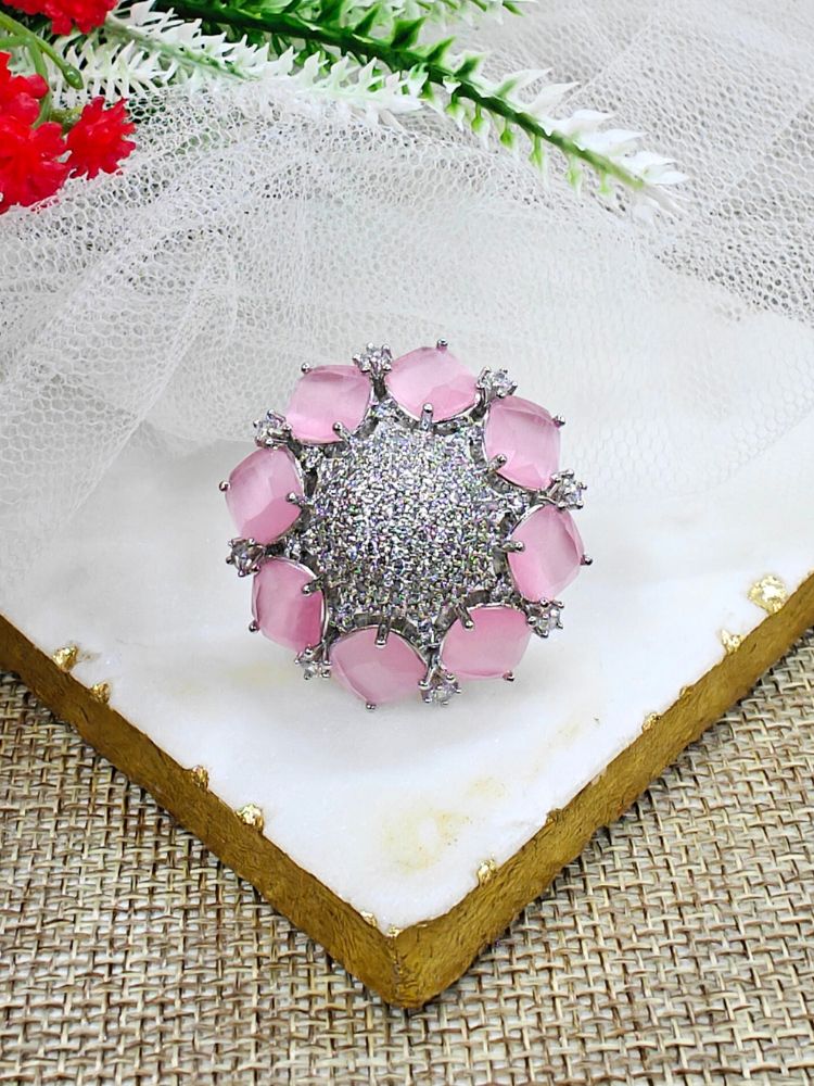 The Mint Pink Aster Ring is a round silver piece with a sparkling, round-shaped center made with white American diamonds, surrounded by petal shaped Mint Pink gemstones, along with an adjustable band. It’s shown on a textured surface with white netting and green leaves in the background.