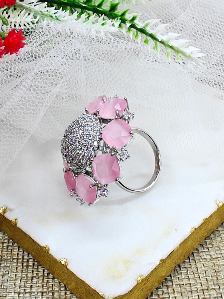 The Mint Pink Aster Ring is a round silver piece with a sparkling, round-shaped center made with white American diamonds, surrounded by petal shaped Mint Pink gemstones, along with an adjustable band. It’s shown on a textured surface with white netting and green leaves in the background.