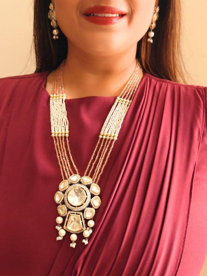 A woman wears the Mint Pink Blair Necklace Set, featuring multi-strand crystals and pearls necklace, along with bog round and square tyani kundan stones, surrounded by american diamonds, along with pearls hanging at the bottom to accentuate the over all look. The necklace comes with matching earrings. She pairs it with a maroon saree, matching earrings, and smiles with red lipstick, her face partially visible.