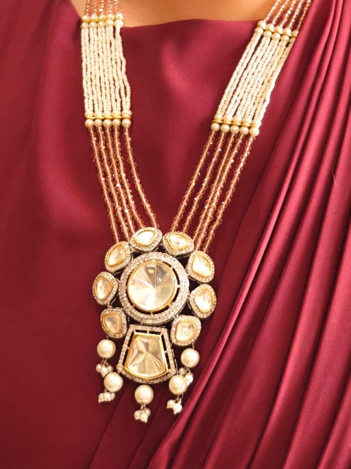 A woman wears the Mint Pink Blair Necklace Set, featuring multi-strand crystals and pearls necklace, along with bog round and square tyani kundan stones, surrounded by american diamonds, along with pearls hanging at the bottom to accentuate the over all look. The necklace comes with matching earrings. She pairs it with a maroon saree, matching earrings, and smiles with red lipstick, her face partially visible.