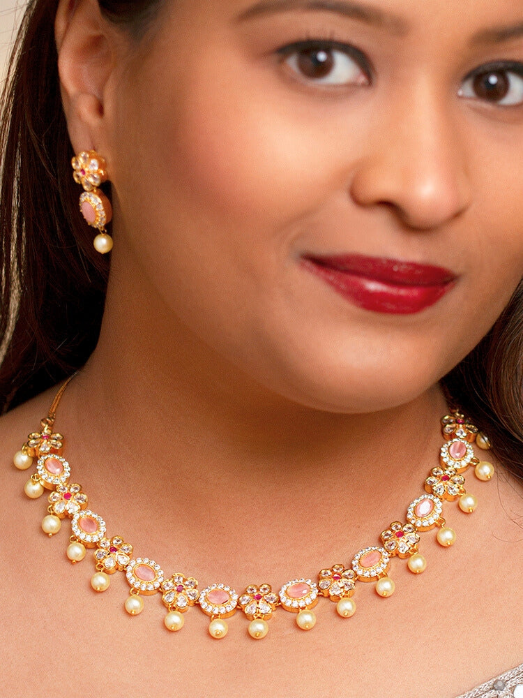 The Mint Pink Samrina Necklace Set entails multiple mint pink stones, surrounded by american diamonds, along with pearls hanging at the bottom.  Comes with matching earrings - is showcased on a black surface, with a patterned cup of crystals and yellow, white, and green artificial flowers in the background.