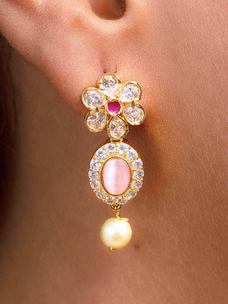 The Mint Pink Samrina Necklace Set entails multiple mint pink stones, surrounded by american diamonds, along with pearls hanging at the bottom.  Comes with matching earrings - is showcased on a black surface, with a patterned cup of crystals and yellow, white, and green artificial flowers in the background.