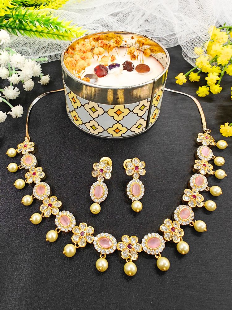 The Mint Pink Samrina Necklace Set entails multiple mint pink stones, surrounded by american diamonds, along with pearls hanging at the bottom.  Comes with matching earrings - is showcased on a black surface, with a patterned cup of crystals and yellow, white, and green artificial flowers in the background.