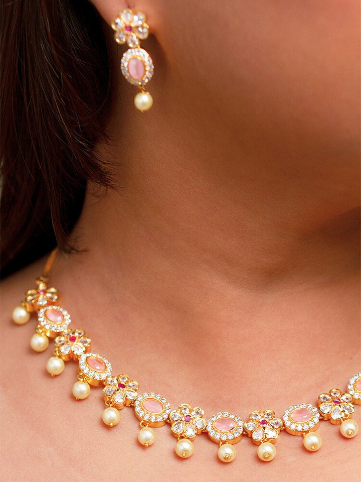 The Mint Pink Samrina Necklace Set entails multiple mint pink stones, surrounded by american diamonds, along with pearls hanging at the bottom.  Comes with matching earrings - is showcased on a black surface, with a patterned cup of crystals and yellow, white, and green artificial flowers in the background.
