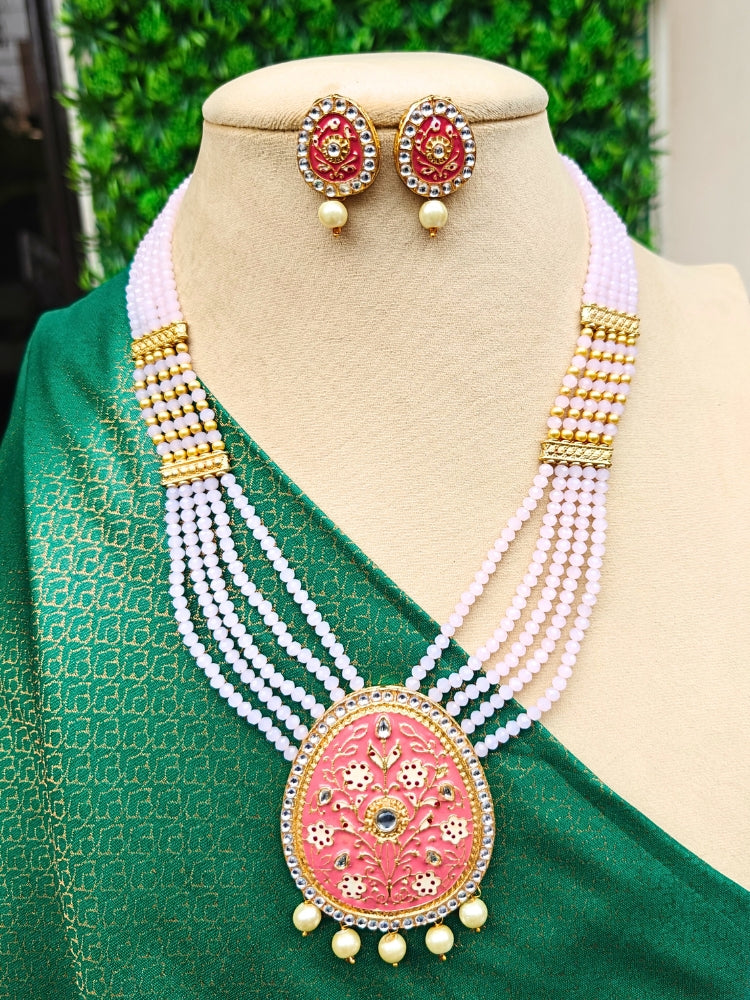 A mannequin showcases the Mint Pink Tulika Necklace Set—brass mint pink crystal bead necklace, with meenakari work pendant, along with matching earrings—paired with a green, gold-patterned saree. Green foliage appears in the background.
