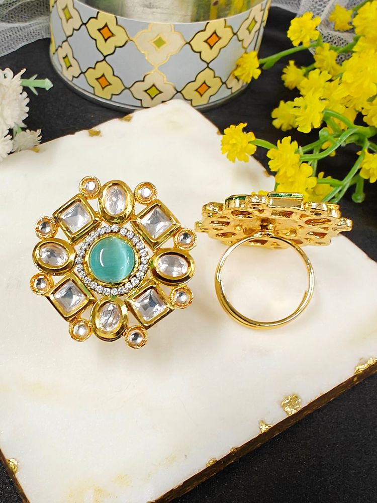 The Mishika Mint Blue Ring, a large ornate gold Tyaani Kundan ring with a central Blue Monalisa Stone and clear gemstones on an adjustable band, is displayed on white marble beside small yellow and white flowers.