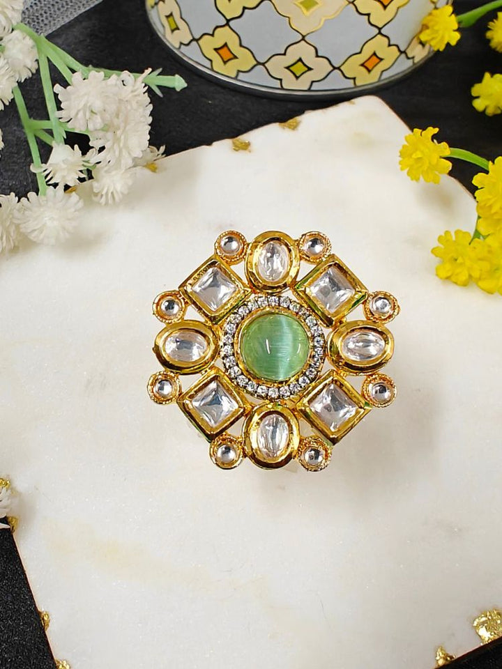 The Mishika Mint Green Ring, a large ornate gold Tyaani Kundan ring with a central Green Monalisa Stone and clear gemstones on an adjustable band, is displayed on white marble beside small yellow and white flowers.