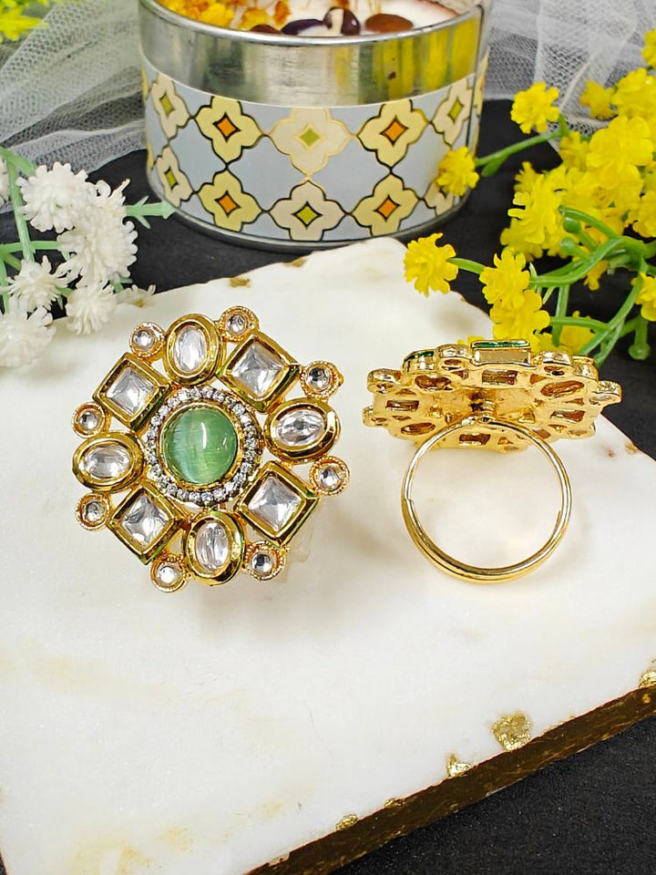 The Mishika Mint Green Ring, a large ornate gold Tyaani Kundan ring with a central Green Monalisa Stone and clear gemstones on an adjustable band, is displayed on white marble beside small yellow and white flowers.