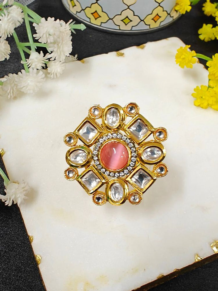 The Mishika Mint Pink Ring, a large ornate gold Tyaani Kundan ring with a central pink Monalisa Stone and clear gemstones on an adjustable band, is displayed on white marble beside small yellow and white flowers.