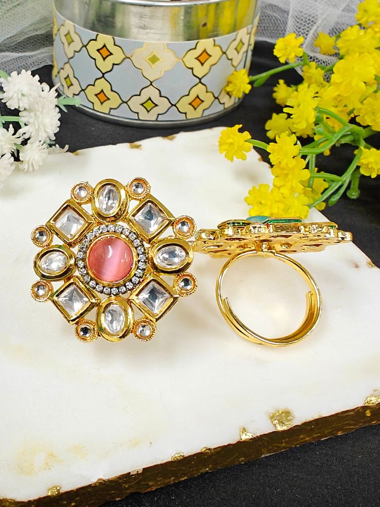The Mishika Mint Pink Ring, a large ornate gold Tyaani Kundan ring with a central pink Monalisa Stone and clear gemstones on an adjustable band, is displayed on white marble beside small yellow and white flowers.