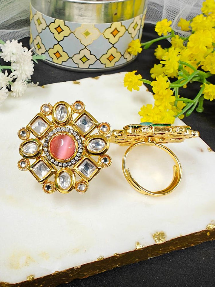 The Mishika Mint Pink Ring, a large ornate gold Tyaani Kundan ring with a central pink Monalisa Stone and clear gemstones on an adjustable band, is displayed on white marble beside small yellow and white flowers.