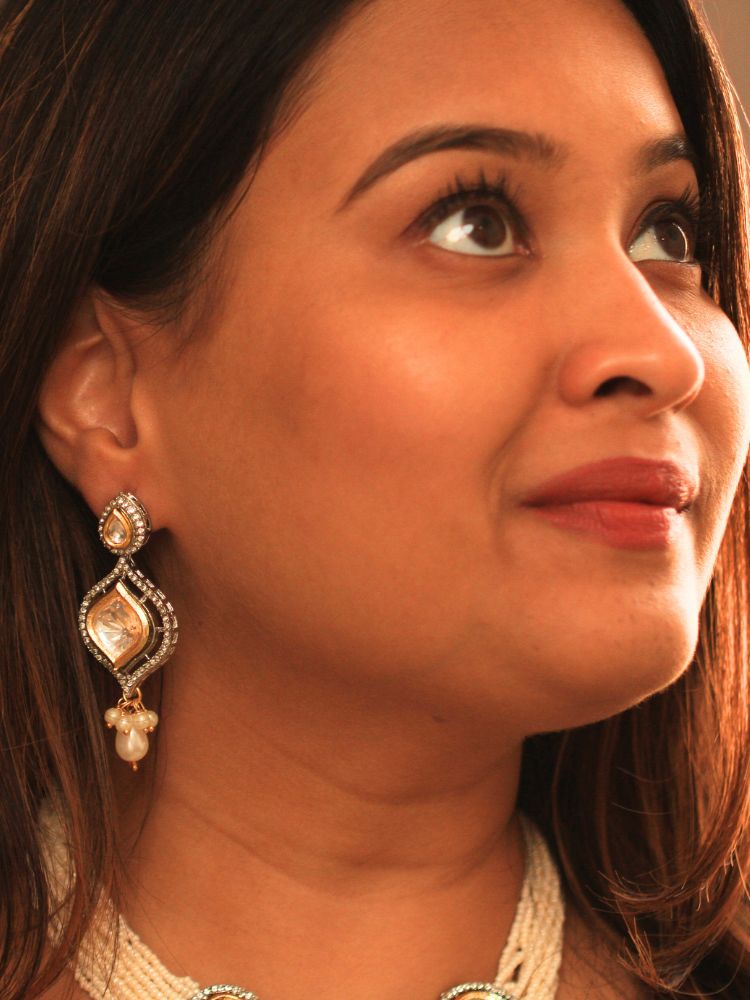 A woman with long brown hair wears the Moissanite & Pearl Reema Necklace Set, featuring multiple strands of pearls, along with kundan moissanite, surrounded by american diamonds, with hanging pearls, along with matching earrings. She smiles softly, her makeup flawless, while the background remains gently blurred. Kundan necklace. Moissanite necklace.