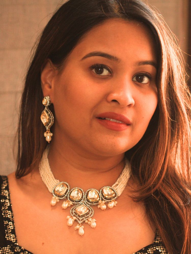 A woman with long brown hair wears the Moissanite & Pearl Reema Necklace Set, featuring multiple strands of pearls, along with kundan moissanite, surrounded by american diamonds, with hanging pearls, along with matching earrings. She smiles softly, her makeup flawless, while the background remains gently blurred. Kundan necklace. Moissanite necklace.