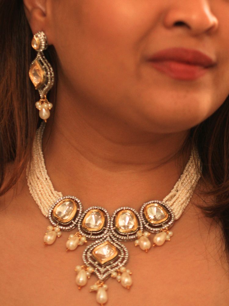 A woman with long brown hair wears the Moissanite & Pearl Reema Necklace Set, featuring multiple strands of pearls, along with kundan moissanite, surrounded by american diamonds, with hanging pearls, along with matching earrings. She smiles softly, her makeup flawless, while the background remains gently blurred. Kundan necklace. Moissanite necklace.