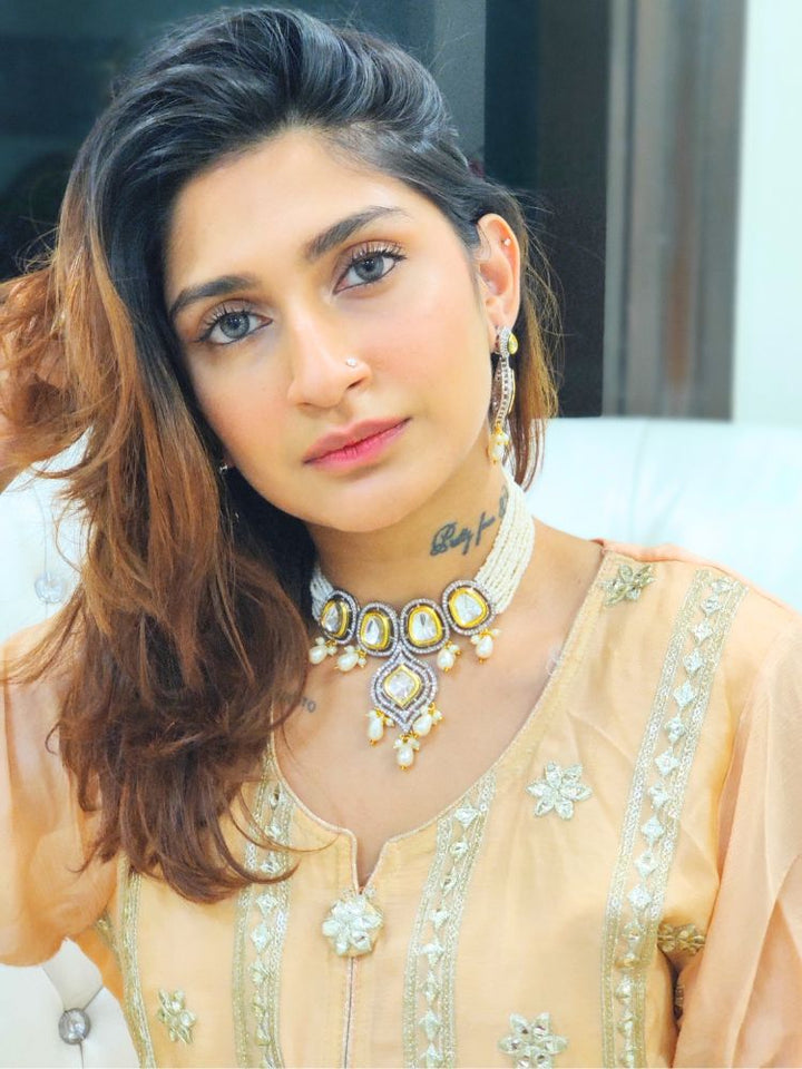 A woman with long brown hair wears the Moissanite & Pearl Reema Necklace Set, featuring multiple strands of pearls, along with kundan moissanite, surrounded by american diamonds, with hanging pearls, along with matching earrings. She smiles softly, her makeup flawless, while the background remains gently blurred. Kundan necklace. Moissanite necklace.