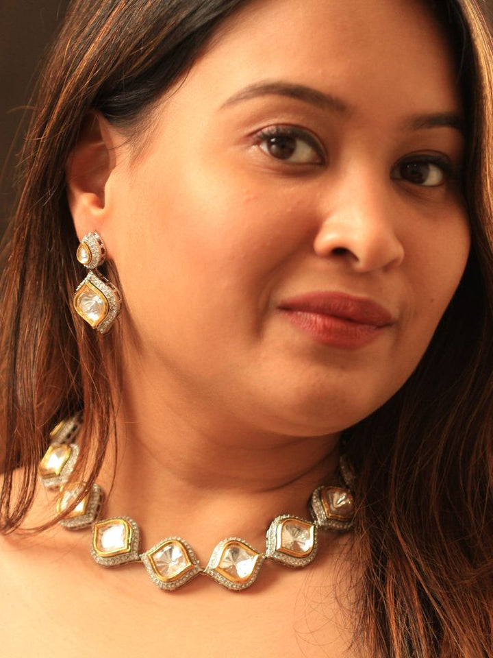 A woman with long brown hair wears the elegant Moissanite Susan Necklace Set, featuring a luxurious necklace and matching geometric earrings all made in kundan, surrounded by American diamonds. With a neutral expression, she glances aside to highlight her beautiful jewelry.