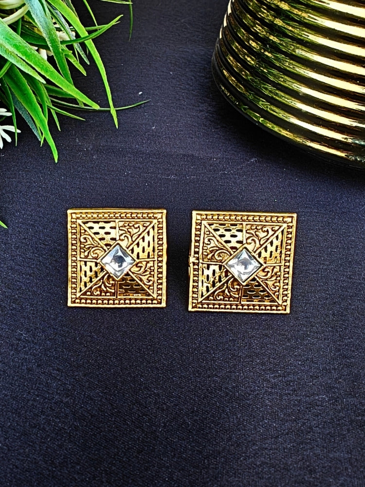 The White Mudita stud Earrings feature square gold filigree with intricate patterns and a White gemstone at the center, elegantly displayed on a black surface beside green leaves and a gold-striped container.