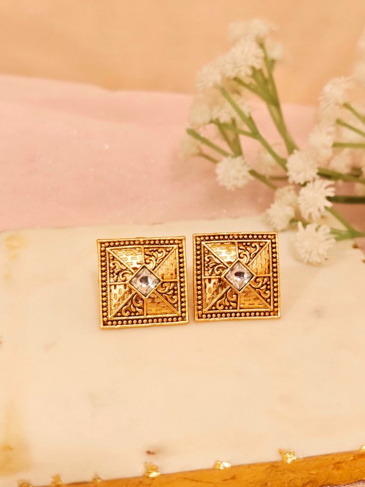 The White Mudita stud Earrings feature square gold filigree with intricate patterns and a White gemstone at the center, elegantly displayed on a White Marble surface beside White Flower and a gold-striped container.