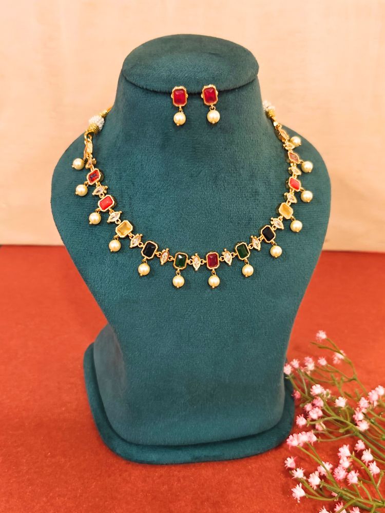 The Multi Color Amelia Choker Necklace Set features colored stones and dangling pearl-like beads, displayed on a teal velvet stand, with matching stud earrings above and pink flowers beside the stand.