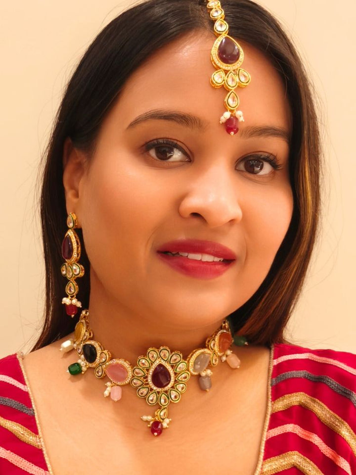 A woman showcases the Multi-Color Aurora Necklace Set, featuring a maang tikka, chandelier earrings, and choker necklace, while dressed in a red and gold outfit against a plain background.