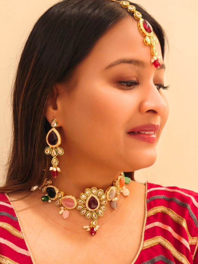 A woman showcases the Multi-Color Aurora Necklace Set, featuring a maang tikka, chandelier earrings, and choker necklace, while dressed in a red and gold outfit against a plain background.
