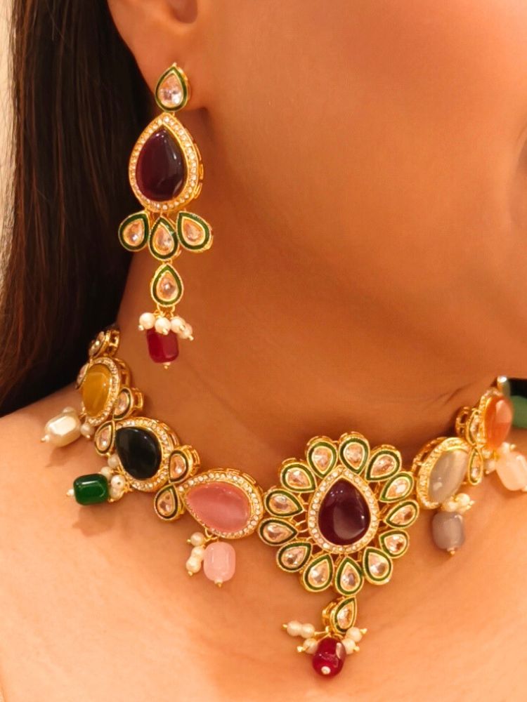 A woman showcases the Multi-Color Aurora Necklace Set, featuring a maang tikka, chandelier earrings, and choker necklace, while dressed in a red and gold outfit against a plain background.
