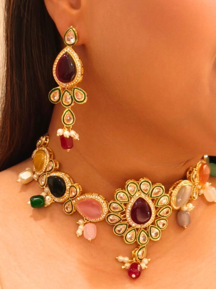 A woman showcases the Multi-Color Aurora Necklace Set, featuring a maang tikka, chandelier earrings, and choker necklace, while dressed in a red and gold outfit against a plain background.